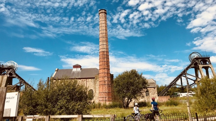 Two children with bikes and cycling gear looking toward old mine building with chimney stack 