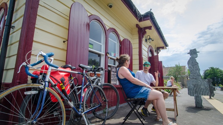Two people sat at table on table outside old fashioned train station waiting room with bikes in the foreground