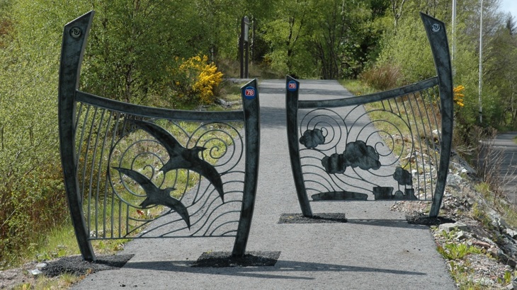 A pair of decorative metal panels, staggered on a traffic-free path to control access. The curved panels have silhouette images on them, one has two birds in flight, the other a puffing steam train. The path beyond continues on uphill where the path enters a wooded area. An interpretation board is in the distance.