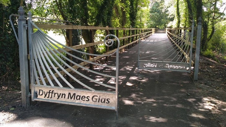 A pair of decorative gates are set at a staggered distance on a traffic-free path. The gates are a pale, galvanized metal. They have sweeping lines which end in a scroll. The words 'Dyffryn Maes Glas' are cut out of the bottom of each gate. The path beyond is long, straight and tree-lined, with a wooden fence running either side.