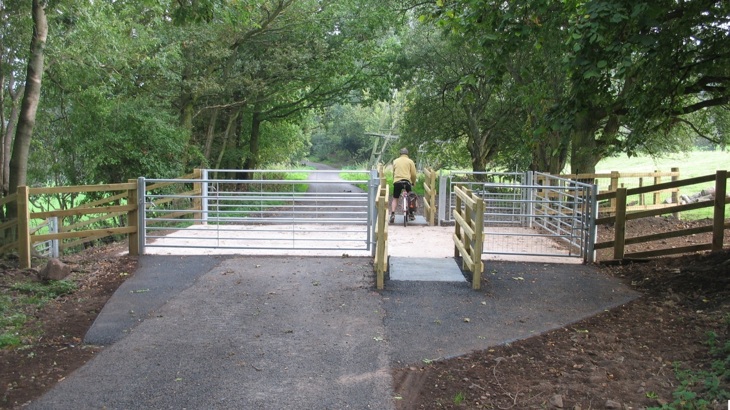 A gated agricultural crossing with cattle grids. In the foreground is tarmac and beyond a series of gates and cattle grids. A person on a bike can be seen from behind, leaving the access point, about to join the main path. It stretches into the distance, is tree-lined and there is a large pylon alongside the path.