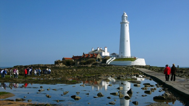 White lighthouse and houses on small island in bay with walkway leading up to it and people walking around it
