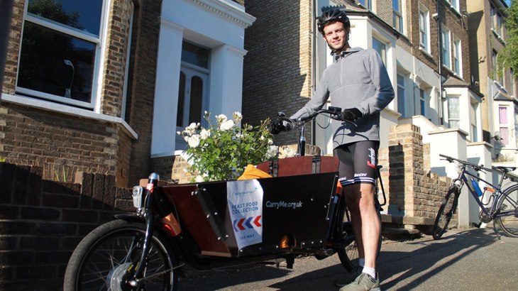 A man stands astride a cargo bike loaded with shopping. He is outdoors on a London street. He is smiling, the weather is fine and the mood is pleasant.