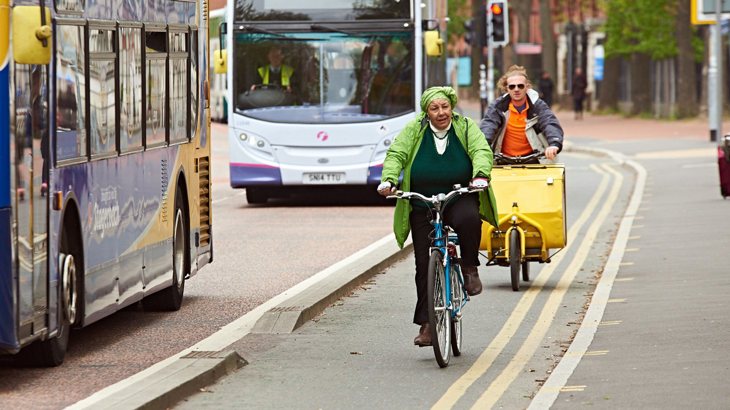 People cycling along a segregated cycling lane next to double-decker buses in a UK city