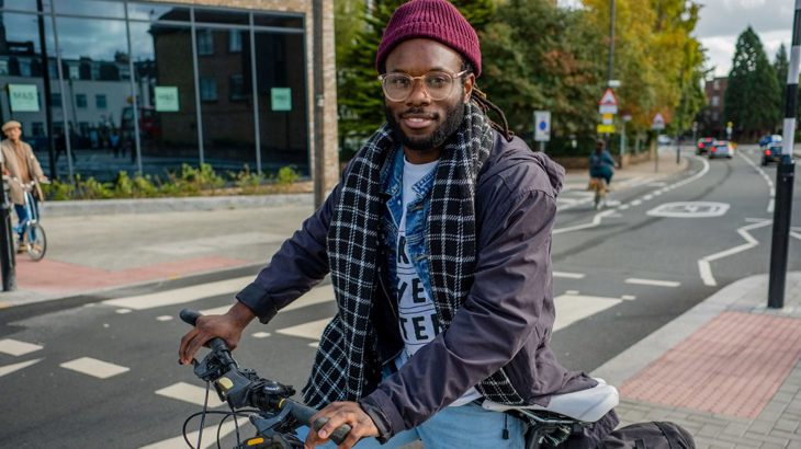 Man wearing a beanie standing with his bicycle smiling at the camera on quiet London street