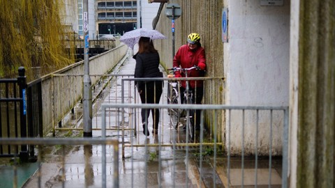 Person on foot with umbrella trying to squeeze past someone on a bicycle through a restrictive barrier on the route