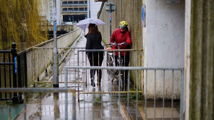 Person on foot with umbrella trying to squeeze past someone on a bicycle through a restrictive barrier on the route