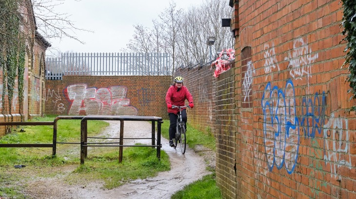 A person cycling past a very small gap next to a barrier on a National Cycle Network route in the rain