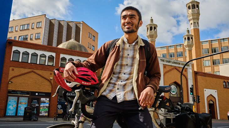 Shaheb stands outside his Mosque with his bicycle and holding his helmet