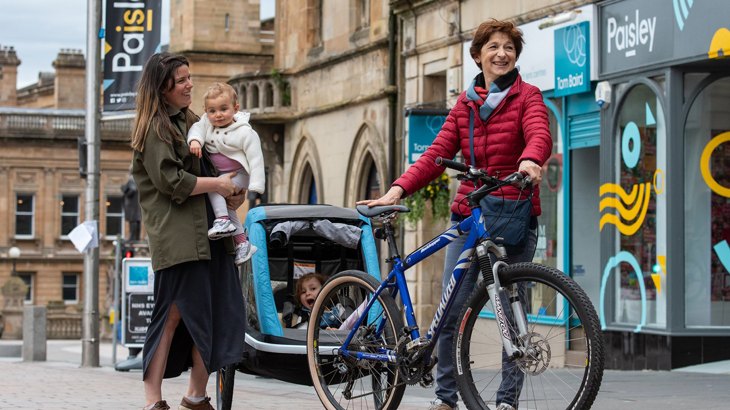 Mum holding baby laughing with friend next to a bicycle with back trailer