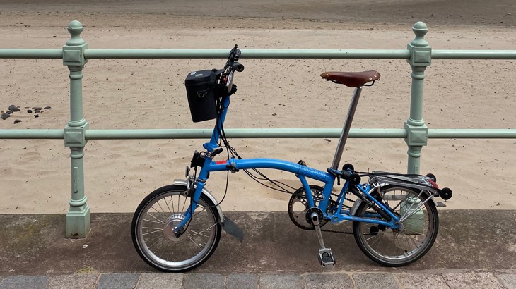 Tierney's blue e-bike propped up against a green railing along the coast with beach in background