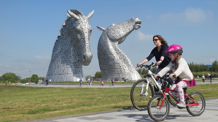 Young girl and woman riding cycles on path near giant horse head sculptures
