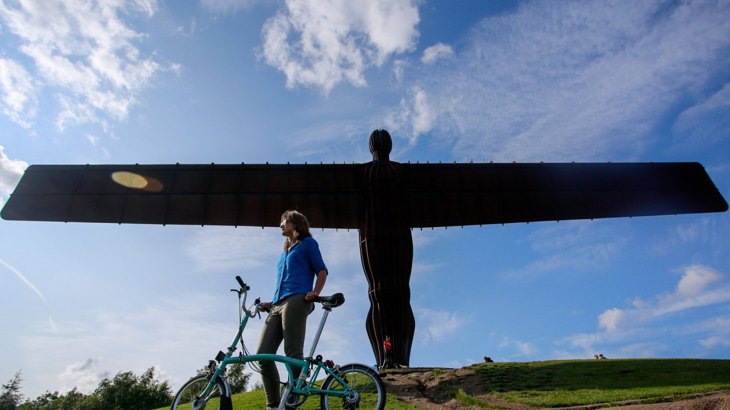 Woman with fold up bike standing in front of large angel sculpture