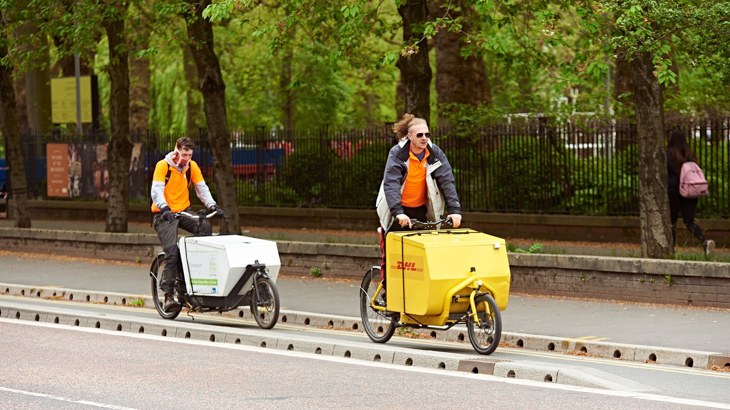 Two men riding cargo bikes along a segregated cycle lane in Manchester, past a tree-lined park.