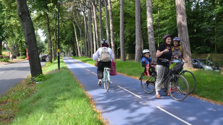 Person cycling a long a blue segregated cycle route lined with green trees as a family with bikes stop and chat to a friend next to the route.