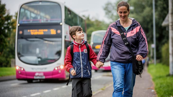 Child looks up at parent as they walk to school along the pavement with buses driving past