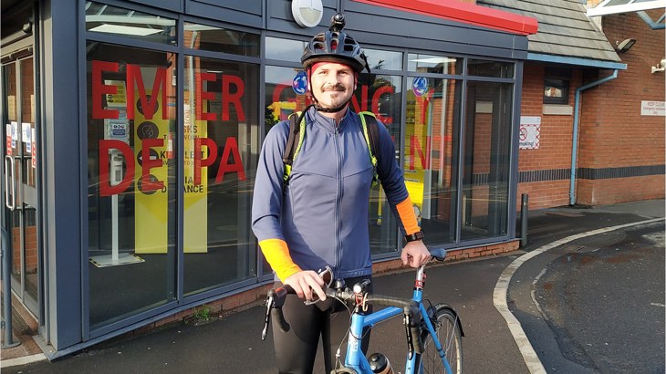 Doctor Matthew Jackson wearing lycra leggings, a waterproof jacket and a helmet, standing with his bike outside his GP surgery
