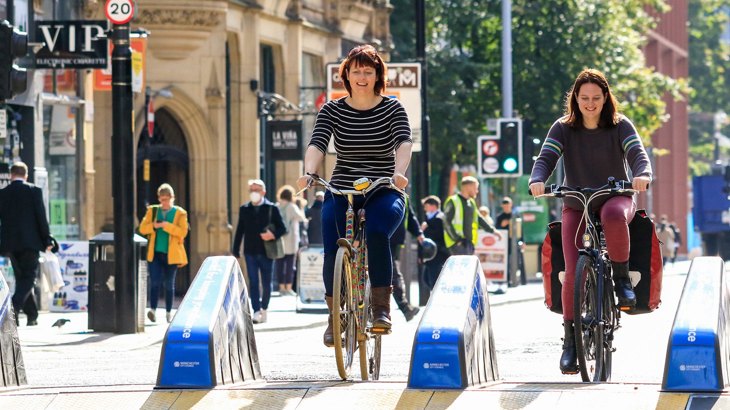 Two women smiling and chatting as they cycle together along a new temporary cycle lane through the high street.
