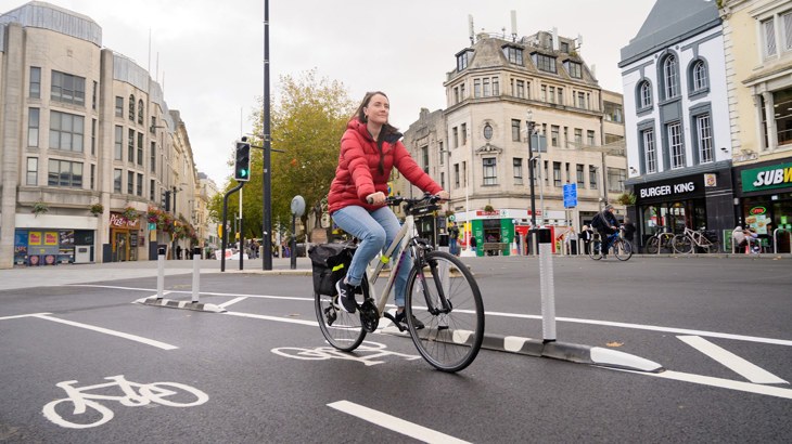 Woman wearing a red waterproof smiling as she cycles along a new temporary cycle lane in a city centre.