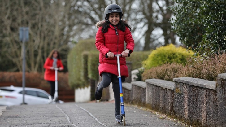 A young girl in school uniform, a winter coat and a helmet rides her scooter.