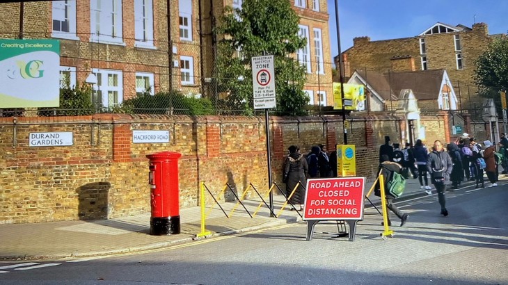 Red sign next to a red postbox, showing that the street outside the school gate is closed to vehicle traffic, as children play and laugh in the street. Healthy Streets Officers London