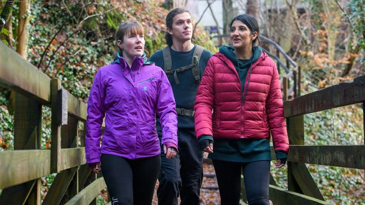 Three friends wearing brightly coloured waterproof jackets walking along a bridge chatting, surrounded by orange autumnal leaves and trees.