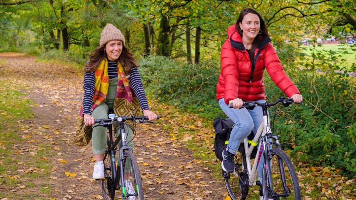 Two women in waterproof jackets and hats cycling through a park surrounded by crisp, orange and brown autumn leaves.