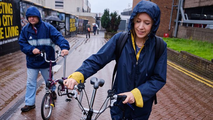 Lady wearing a navy blue waterproof smiling as she walks down a street with her bike in the rain.