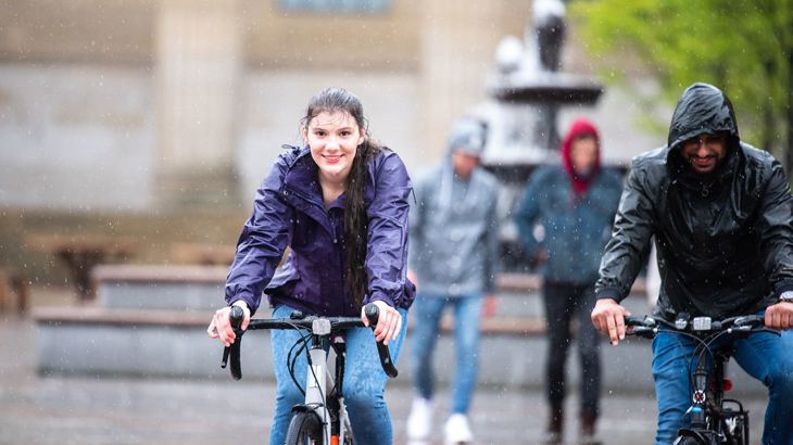 Lady wearing a purple waterproof smiling as she cycles in the pouring down rain next to her male friend in a black waterproof jacket with his hood up.