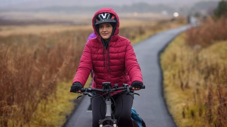 Lady wearing a red coat with the hood pulled up over her cycle helmet, cycling down a traffic-free path with grey skies in the background.