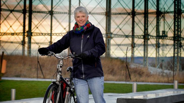 Lady wrapped up in a black fleece coat, scarf and gloves stands smiling with her bicycle next to her.
