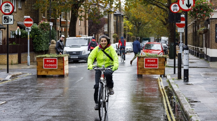A lady rides her bike on a road with no moving cars. There are two planters behind sayin 'road closed'. It is a wet, autumn day. 