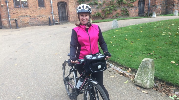 Caroline wearing a black and pink waterproof jacket, black trousers and her cycle helmet, standing with her bicycle.