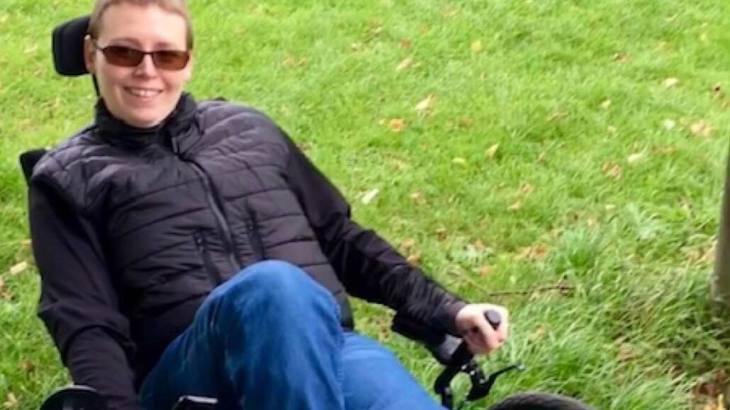 Kelly, wearing a black jacket and blue jeans, sitting on her ICE trike - an adapted bicycle - smiling at the camera.