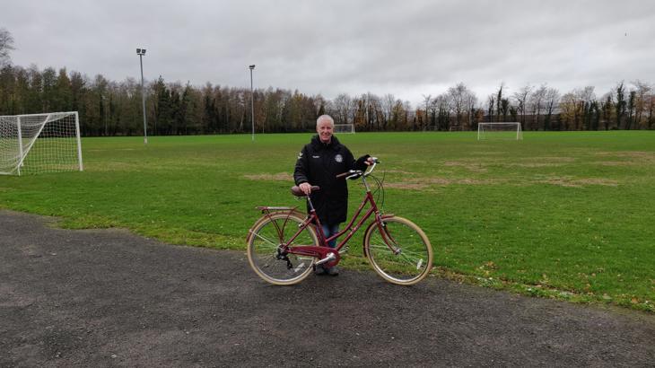 Jane, white female, stands with her maroon Dutch-style cycle on a gravel path on the edge of a sports field. Jane has cropped white hair and is wearing a black coat and blue jeans. There are several football posts in the field behind her, and trees in the distance.