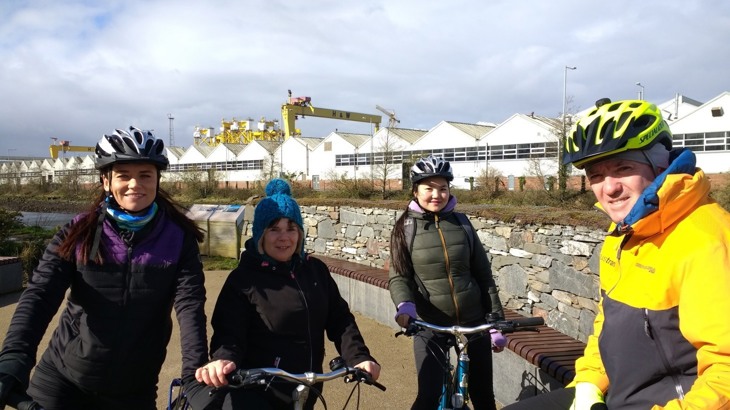 three women on a group ride with Walk Wheel Cycle Trust staff member Tom in a park with Belfast Harland and Wolf cranes in the background