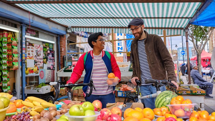 A man and woman holding their bikes, chatting as they buy fruit at a food market in London.