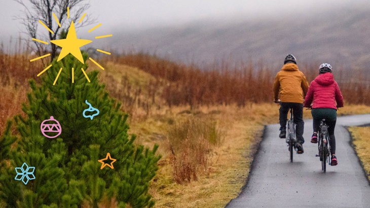 Two people cycling on a traffic-free path past a Christmas tree with illustrated decorations