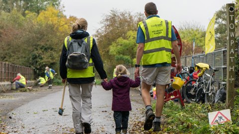 Mum and dad holding hands with their small daughter, wearing Walk Wheel Cycle Trust volunteer high-vis jackets and walking away from camera down a traffic-free route.