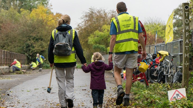 Mum and dad holding hands with their small daughter, wearing Walk Wheel Cycle Trust volunteer high-vis jackets and walking away from camera down a traffic-free route.