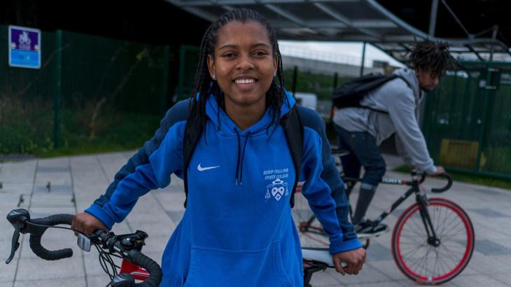 Girl wearing a hoodie stands with her bicycle smiling at the camera whilst a man cycles past in the background.
