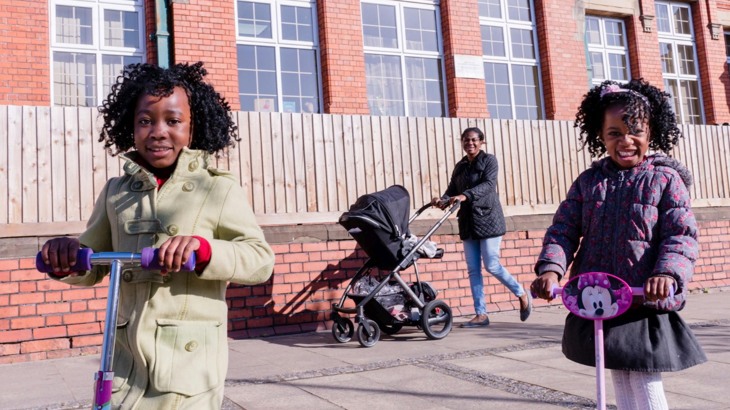Two young girls on their scooters smile at the camera. Their mum is walking behind with a pushchair