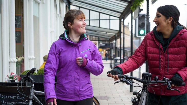 Two women wearing brightly coloured raincoats, chatting and walking with their bikes through a high street
