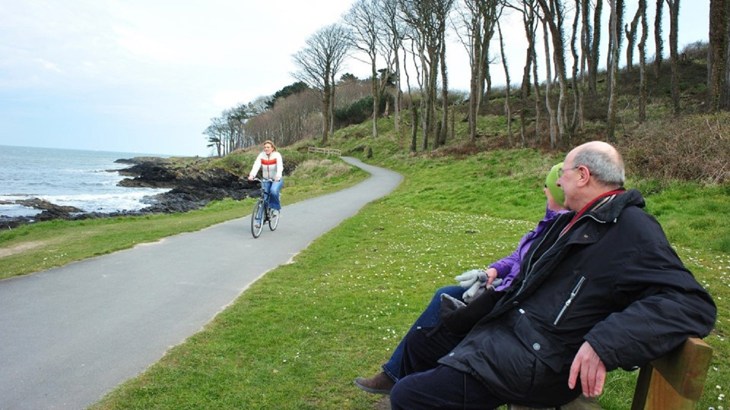 A cyclist on the North Down Coastal Path watched by two people sitting on a bench 