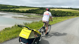 A man wearing a white t-shirt cycling along The Camel Trail with a trailer and two young children sat inside on a sunny day