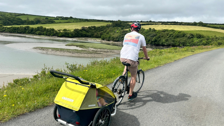 A man wearing a white t-shirt cycling along The Camel Trail with a trailer and two young children sat inside on a sunny day
