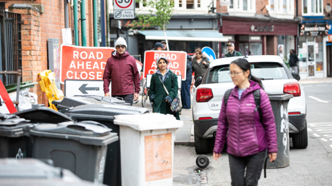 A sidewalk lined with bins, road signs and cars parked, with pedestrians navigating their way around them.