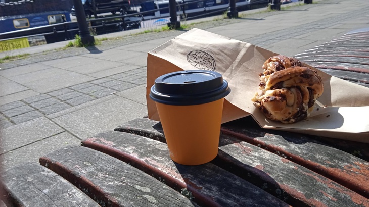 A coffee and baked pastry on a bench with a view of a canal with river boats on it.