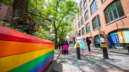 Two people in Manchester city centre next to a pride flag one person has short hair and is in a wheelchair and the other is walking alongside wearing a tutu 