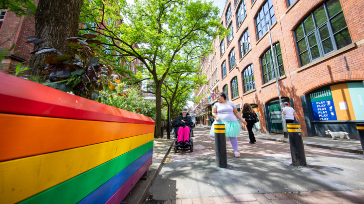 Two people in Manchester city centre next to a pride flag one person has short hair and is in a wheelchair and the other is walking alongside wearing a tutu 